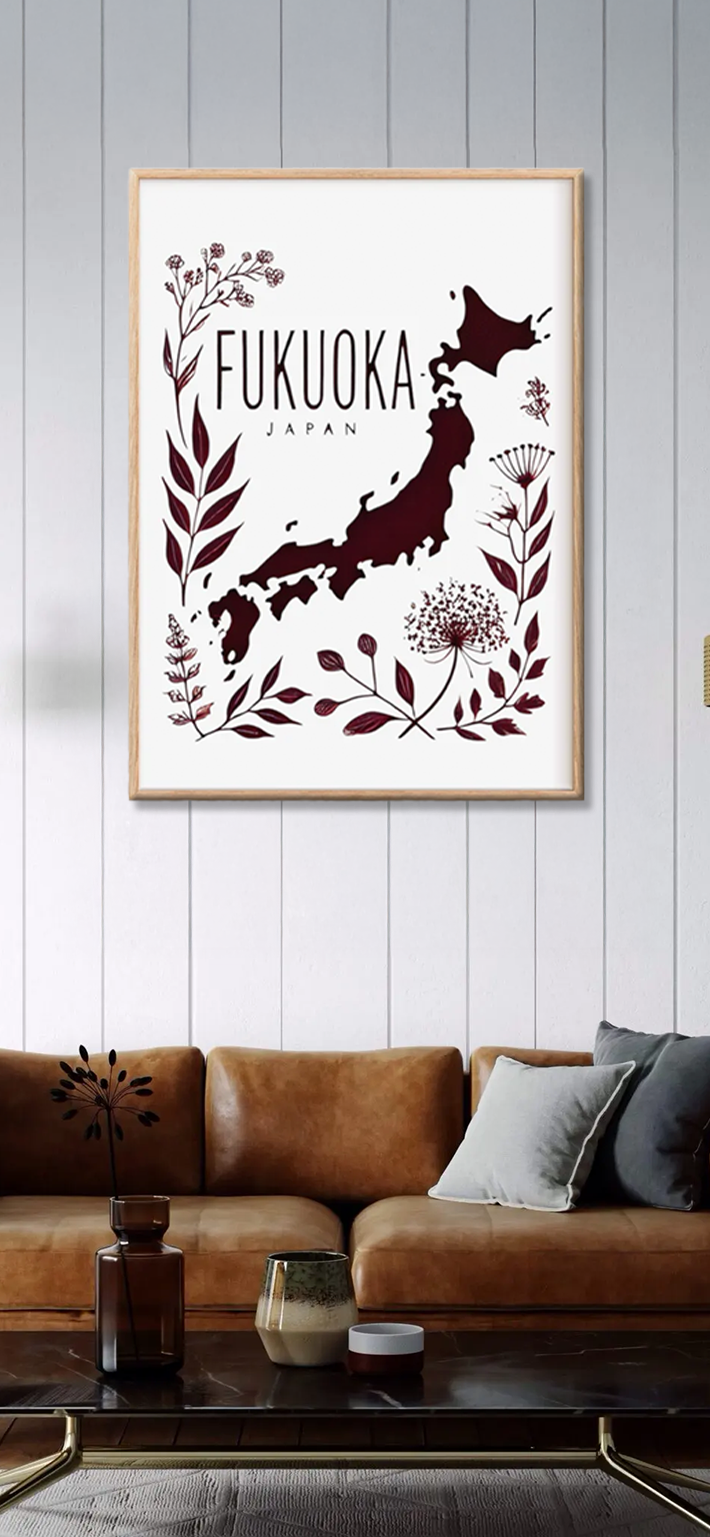 A framed wall art print of Japans map with Fukuoka Japan written above, surrounded by maroon botanical illustrations, hangs above a brown leather sofa with gray and blue cushions in a modern living room.